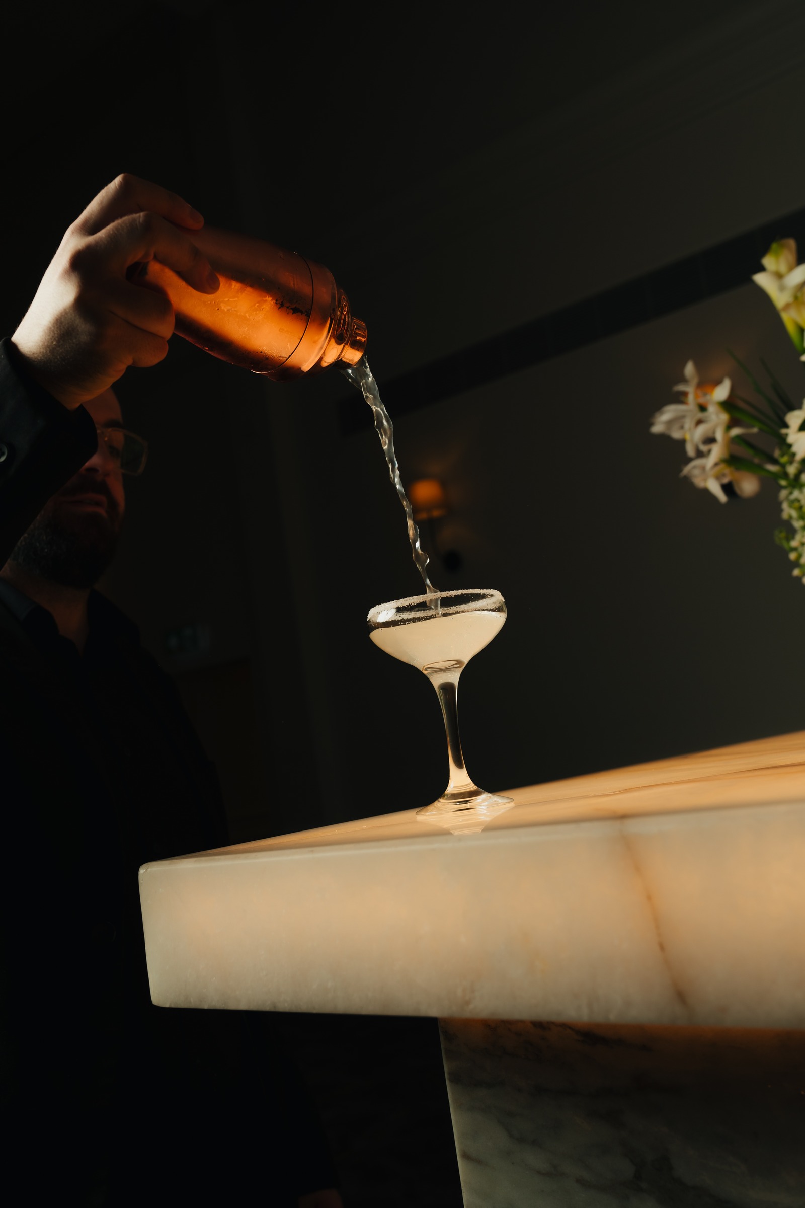 Bartender pouring a cocktail into a coupe glass on the illuminated marble bar