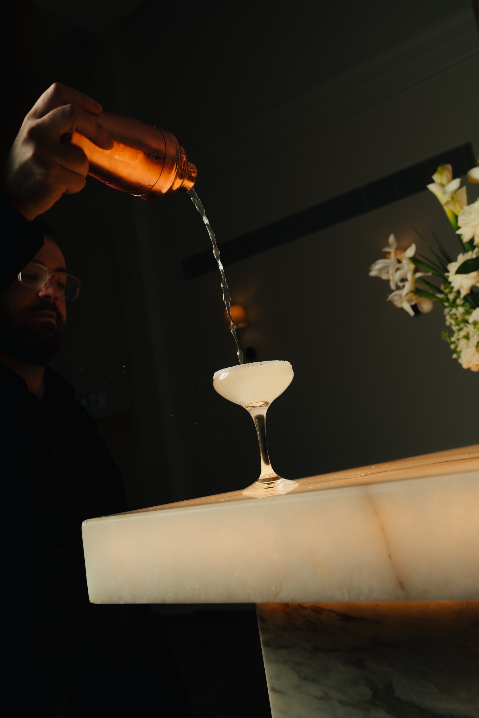 Cocktail being poured into a coupe glass on the backlit onyx bar at Melrose