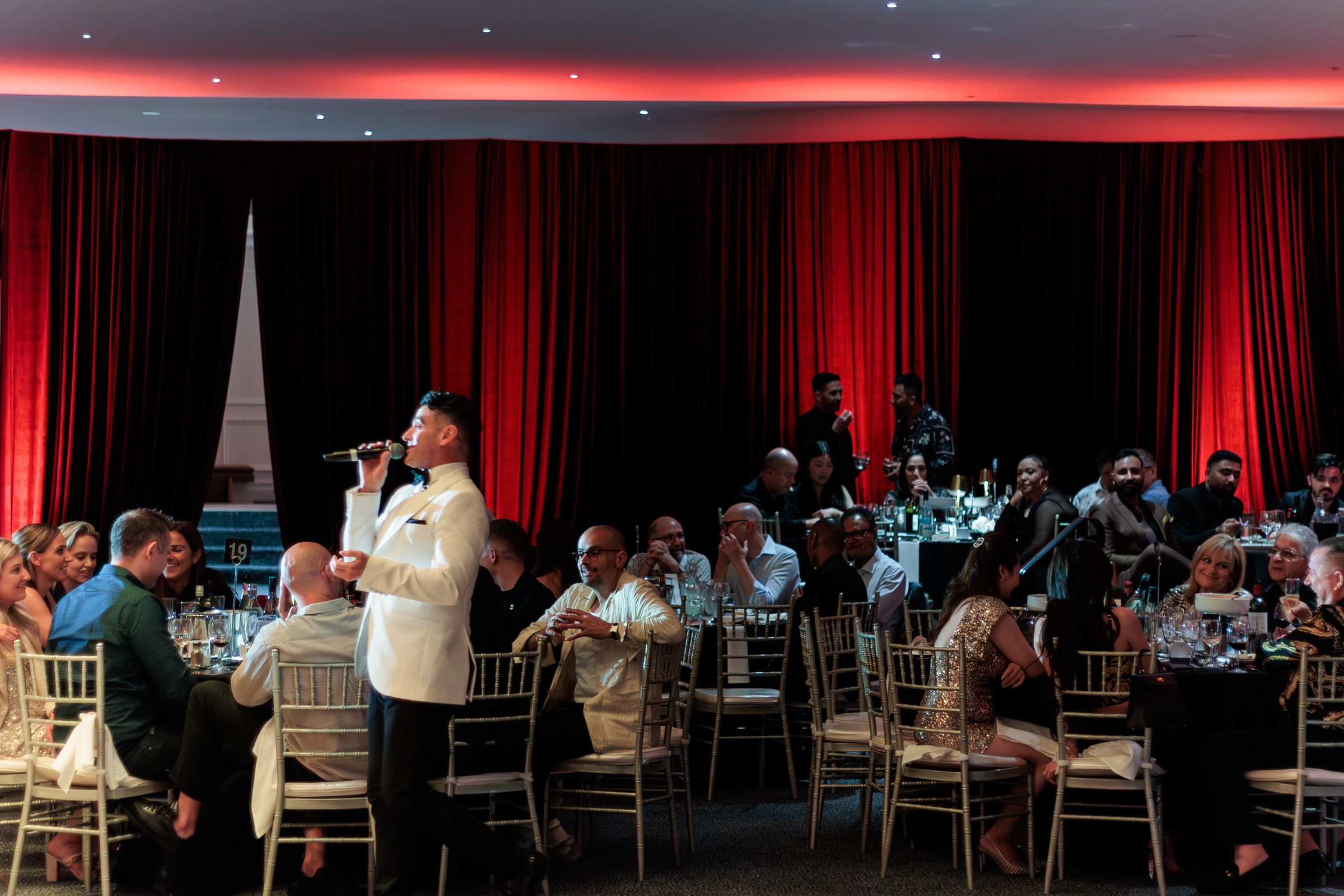 Singer in a white jacket performing to seated guests in front of red drapes