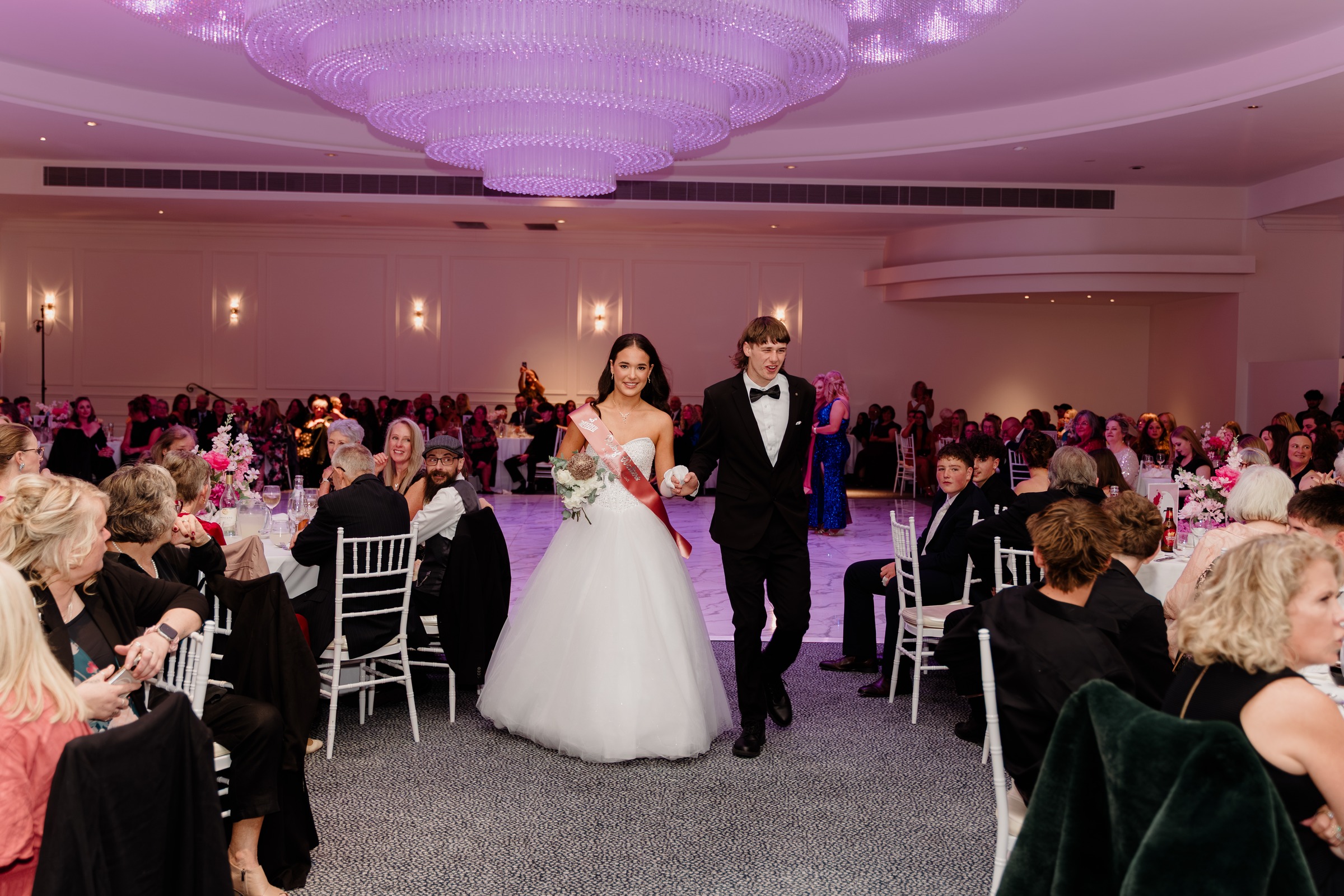 Debutante couple walking between the tables beneath the chandelier at Melrose Events