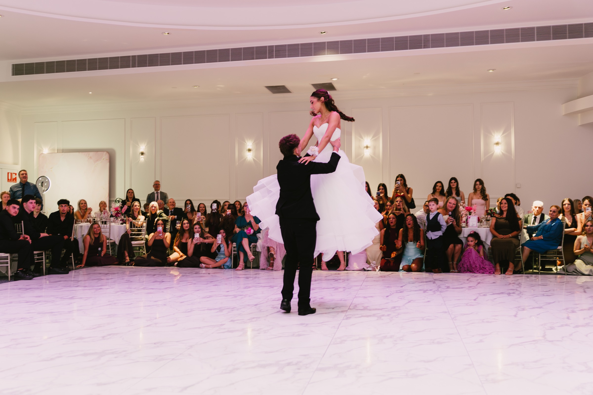 Debutante lifted high by her partner on the marble dance floor as guests watch