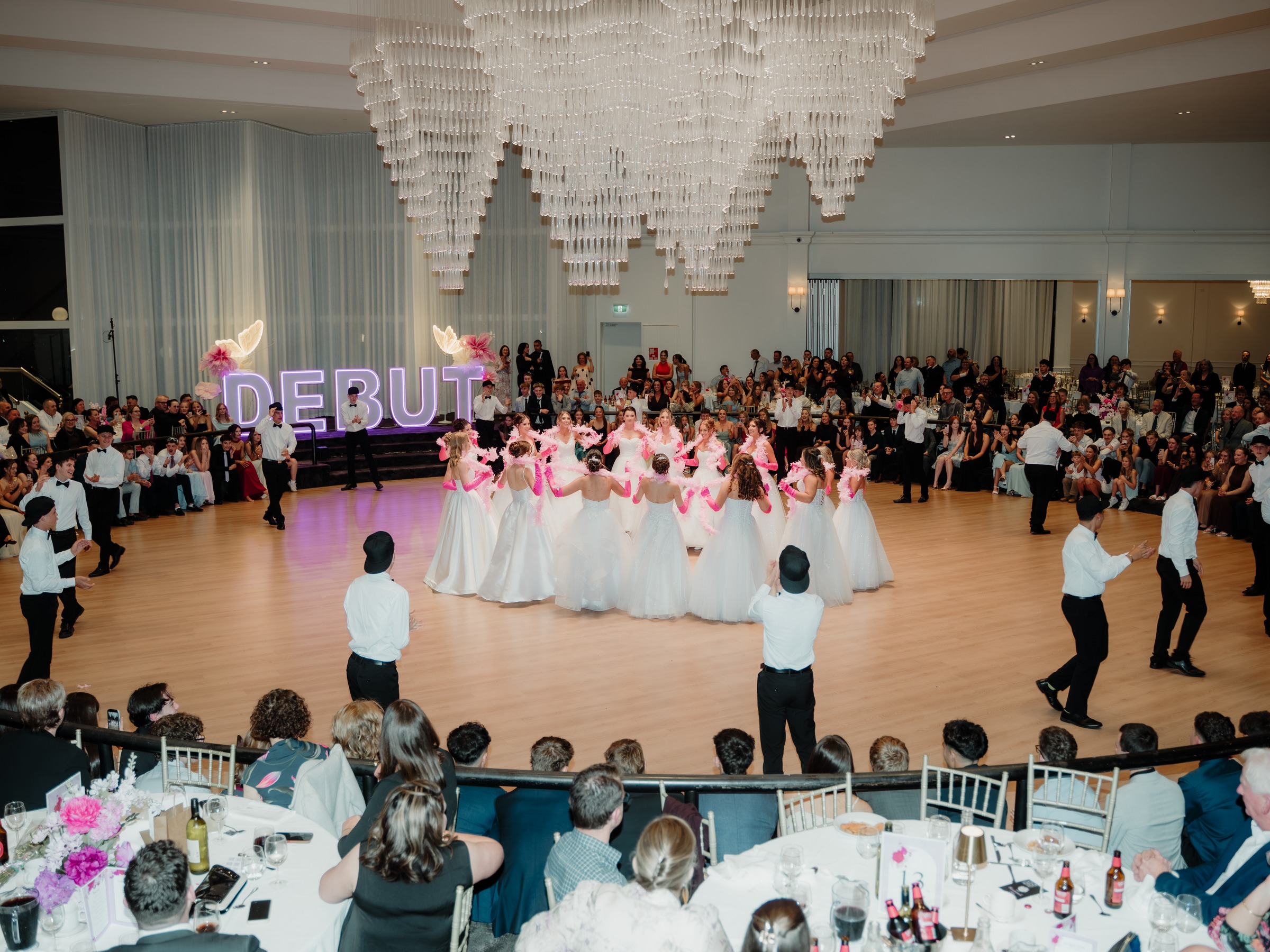 Debutantes in white gowns gathered before the Debut sign with a full ballroom of guests