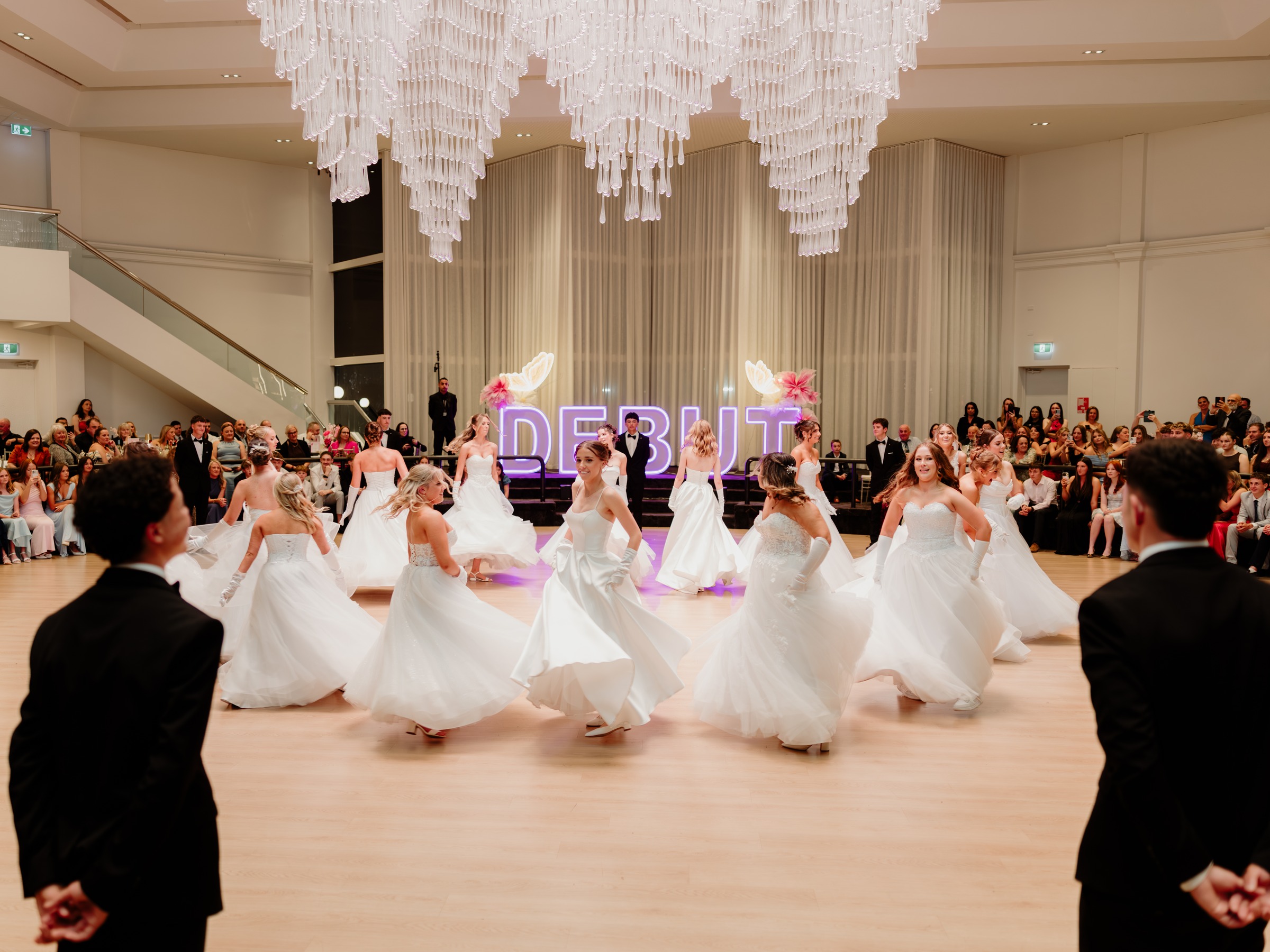 Debutantes in white gowns dancing in a circle under the chandelier at Melrose Events