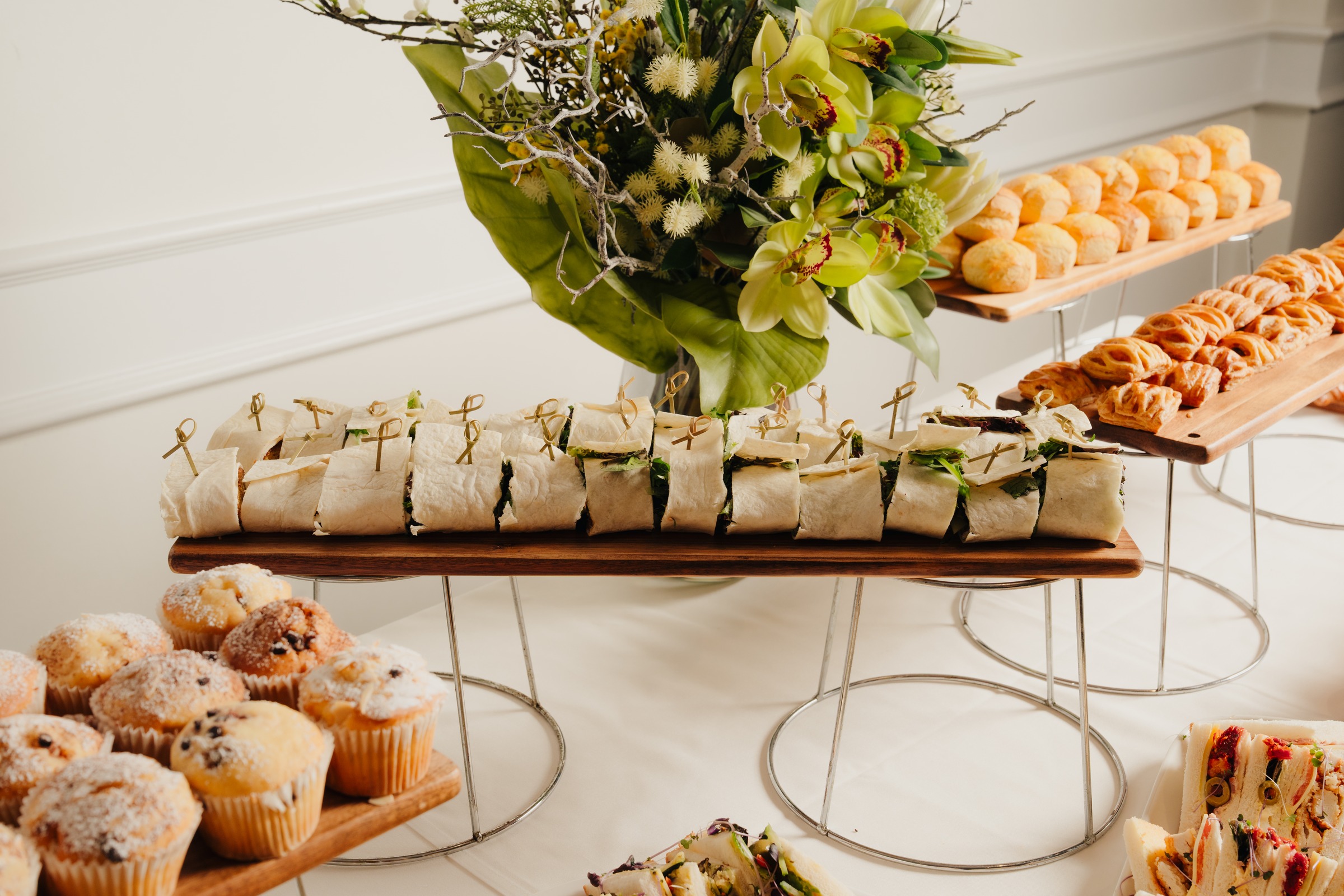 Morning tea grazing table with wraps, muffins and pastries under floral arrangement