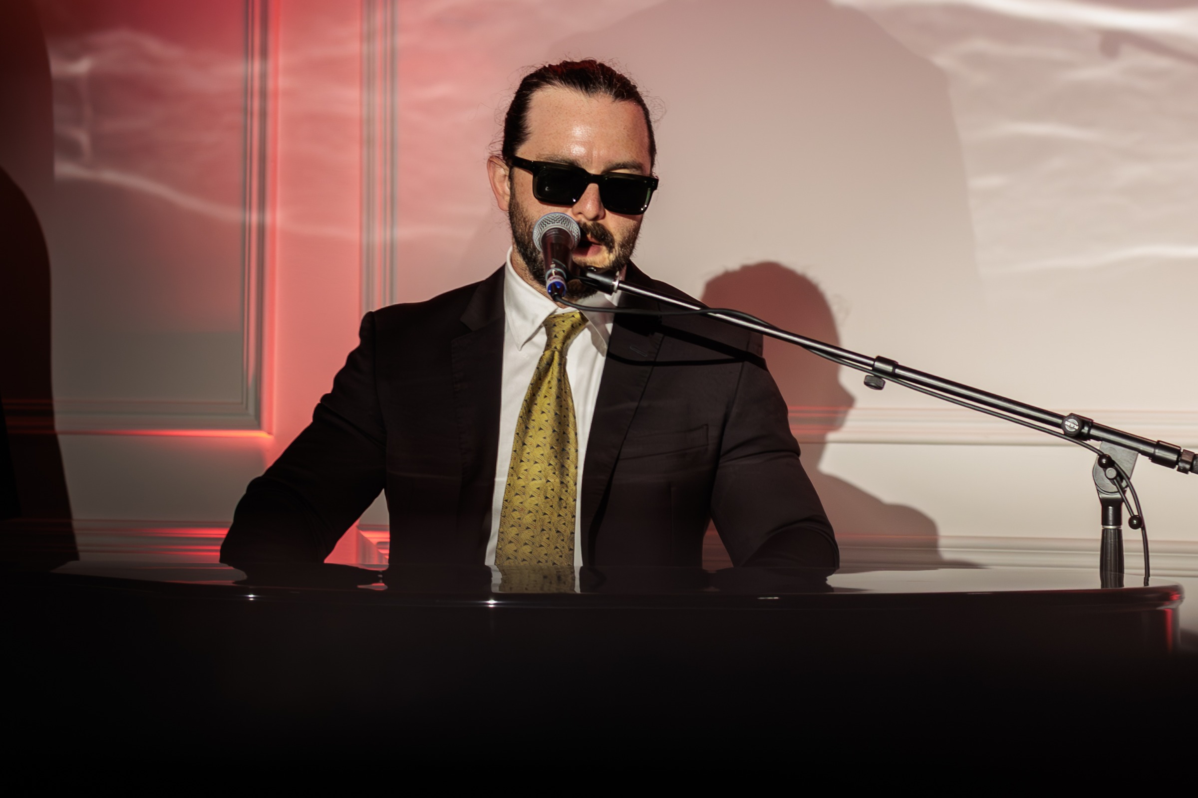 Pianist in sunglasses and suit singing at a grand piano under red light